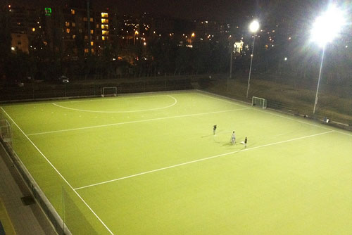 A well-maintained field hockey pitch, with neatly trimmed green turf and white boundary lines marking the playing area. The surrounding areas are also well-manicured, with tall trees visible in the background.